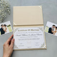 Load image into Gallery viewer, A hand is holding a gold-colored wedding certificate with the names of the newlyweds, Sarah Williams and James Brown, on a gray surface.
