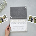 Load image into Gallery viewer, A hand is holding a white wedding certificate with gold lettering, which appears to be a marriage certificate. The certificate is placed on a gray surface, surrounded by two photographs of a couple.
