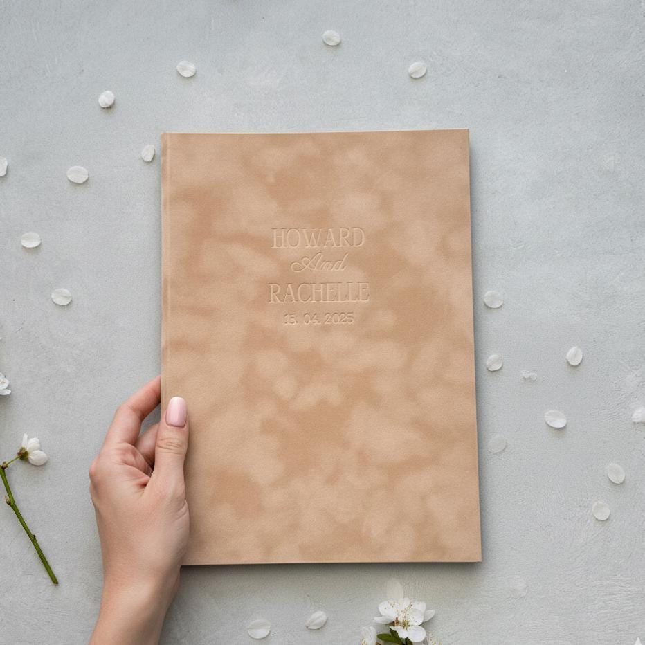 A hand is holding a book titled &quot;Howard and Rachelle&quot; with the date &quot;15.05.2025&quot; printed on it, surrounded by scattered white flower petals.
