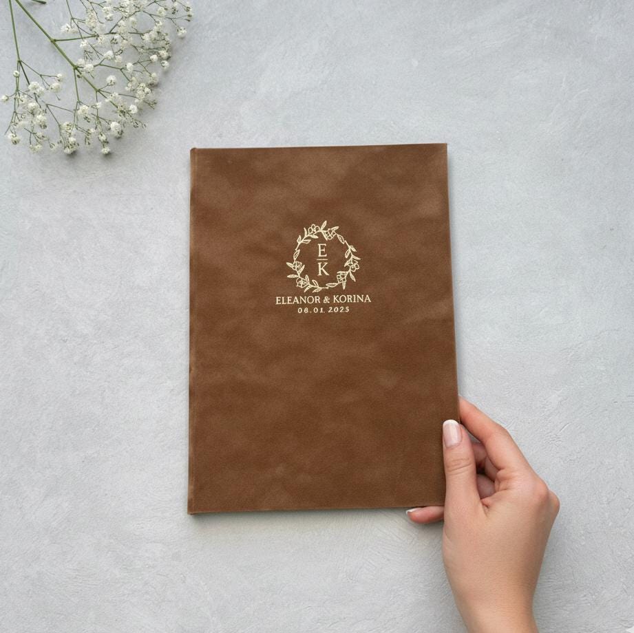 A hand is holding a brown leather-bound book with a gold embossed monogram and text on the cover, which appears to be a wedding invitation or guest book.