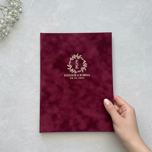A hand is holding a burgundy-colored book with a gold embossed monogram and text on the cover.