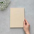 Load image into Gallery viewer, A hand is holding a cream-colored book with the text "HOWARD & RACHELLE" printed on it, placed on a gray surface next to a small bouquet of white flowers.
