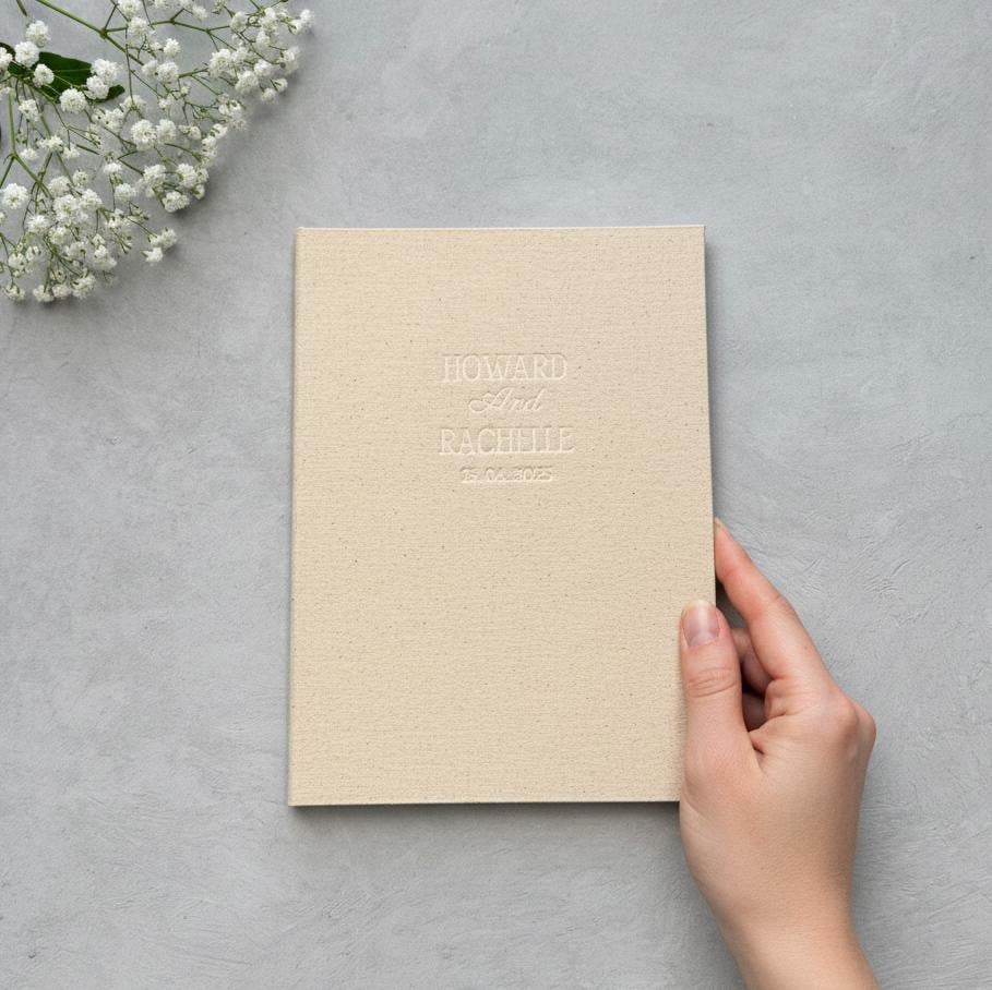 A hand is holding a cream-colored book with the text &quot;HOWARD & RACHELLE&quot; printed on it, placed on a gray surface next to a small bouquet of white flowers.